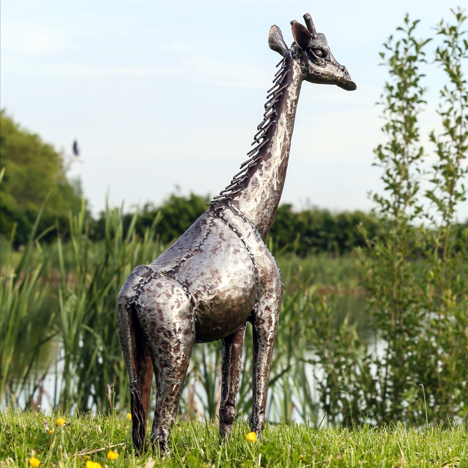 Metallic giraffe sculpture in a natural setting with greenery and water in the background